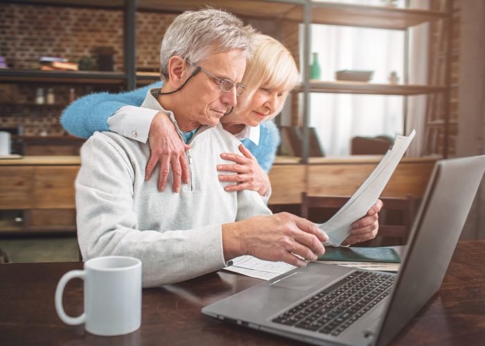 A view from the angle. Man is working and studying information from papers. He works at the table. She is standing behand him and huggings him. Also old woman is reading a document with her husband